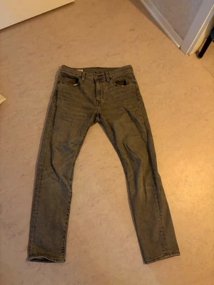 Levis 502  - Säljer ett par grå jeans med klassisk femficksmodell och raka ben. Jeansen har bälteshällor, knappgylf och en liten röd detalj på bakfickan. Materialet är denim i bomull och passformen är normal. Perfekta till en avslappnad stil.