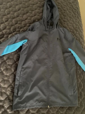 Grå vindjacka med blå detaljer - Snygg grå vindjacka från Under Armour med blå detaljer längs ärmarna. Jackan har huva, hel dragkedja framtill och meshfoder på insidan för extra komfort. Perfekt för blåsiga dagar och sportiga outfits.