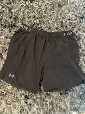 Svarta träningsshorts Under Armour - Svarta träningsshorts från Under Armour med elastisk midja och logga på benet samt linningen. Shortsen har fickor på sidorna och är tillverkade i ett lätt och ventilerande material, perfekta för träning eller sport.