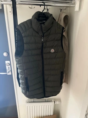 Olivgrön dunväst från Moncler - Snygg olivgrön dunväst från Moncler med hög krage och tvåvägsdragkedja framtill. Västen har två fickor med dragkedja och Moncler-logga på bröstet. Perfekt för lager-på-lager och håller dig varm under kyliga dagar.
