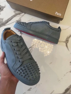 Gråa Louboutin - Säljer ett par gråa Christian Louboutin i mocka med nitar på tån och längs sidorna. Skorna har klassisk röd sula, rund tå och matchande grå skosnören. Insidan är beige och ger en lyxig känsla. Perfekta för dig som vill sticka ut.