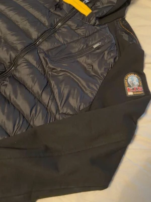 Svart dunväst från Parajumpers L - Snygg svart dunväst från Parajumpers med gul dragkedjedetalj och patch på axeln. Västen har en bröstficka med dragkedja och orange innerfoder. Perfekt för lager-på-lager och streetwear-vibe.