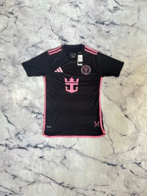 Svart fotbollströja med rosa detaljer - Snygg svart fotbollströja från Adidas med rosa detaljer och tryck. Tröjan har korta ärmar, rund hals och klubbmärke samt sponsorlogga på bröstet. På ryggen står det 'Messi' och nummer 10 i rosa. Perfekt för dig som älskar fotboll och vill sticka ut på planen.