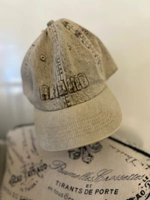 Beige denimkeps med broderi - Säljer en beige keps i denim med böjd skärm och broderad text framtill. Kepsen har en avslappnad vintage-look och metallöljetter för ventilation. Passar perfekt till en streetwear-stil och är enkel att matcha med olika outfits.