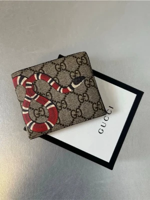 Gucci plånbok med orm-motiv - Cool plånbok från Gucci i beige canvas med klassiskt GG-mönster och en röd, vit och svart orm tryckt på framsidan. Plånboken har en fyrkantig form och känns lyxig med sin ikoniska design. Perfekt accessoar för dig som gillar statement-detaljer.