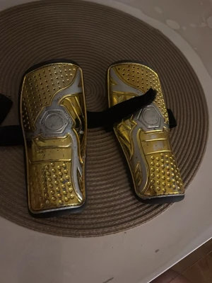 Guldiga benskydd med svart rem - Säljer ett par sportiga benskydd i guld och silver med svart kardborreband. Skydden har en cool, metallisk look och är tillverkade i plast med vadderad insida för extra komfort. Perfekta för fotbollsträning eller match!