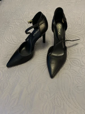 Svarta pumps med ankelrem från Catwalk - Stilrena svarta pumps med spetsig tå och smal klack från Catwalk. Skorna har en elegant ankelrem med guldfärgat spänne och är tillverkade i ett slätt material som ger en klassisk look. 