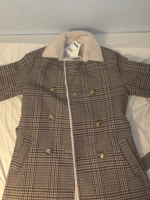 Kapa från C’est Beau La Vie - Snygg rutig kappa i brunt, beige och mörkblått med dubbelknäppning och stora guldknappar. Kragen är i mjuk fuskpäls och kappan har två fickor framtill. Perfekt för dig som vill ha en klassisk men trendig look.