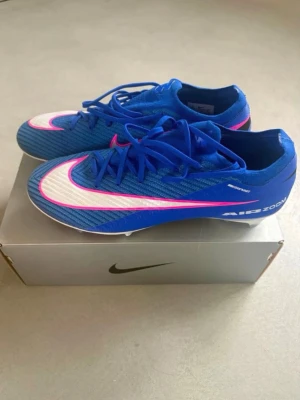 NIKE AIR ZOOM MERCURIAL fotbollsskor  - Snygga Nike Air Zoom fotbollsskor i blått med vita och rosa detaljer. Skorna har en låg profil, snörning och mönstrad yttersula för bra grepp på planen. Tillverkade i syntetmaterial med tydlig Nike-logga och Air Zoom-tryck på sidan.