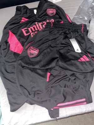Arsenal tracksuit  - Säljer ett Arsenal träningsset från Adidas i svart med rosa detaljer. Setet består av en långärmad tröja med halv dragkedja och matchande byxor. Båda plaggen har Arsenal-logga och Adidas-märke samt rosa ränder och paneler. Perfekt för fotbollsträning eller gym.