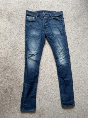 Blå slim fit jeans med slitningar från Diesel  - Snygga blå slim fit jeans med slitningar och tvättade detaljer på låren och knäna ifrån Diesel. Klassisk med knappgylf och bälteshällor. Jeansen har en modern passform och är perfekta för en avslappnad stil. Använda ej av mig längre. 