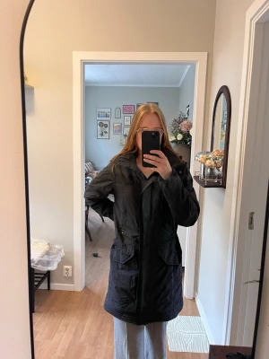 Svart parkas från Tenson - Snygg svart parkas från Tenson med hög krage, dragkedja och tryckknappar framtill. Jackan har stora fickor med lock och justerbar midja. Tyvärr har jag behövt laga den lite se bild. Den är nytvättad 