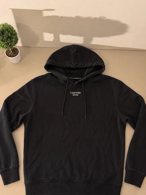 Svart hoodie från Calvin Klein Jeans - Snygg svart hoodie från Calvin Klein Jeans med vit logga broderad på bröstet. Klassisk modell med huva och dragsnören samt ribbade muddar vid ärmar och nederkant. Perfekt för en chill och stilren look.