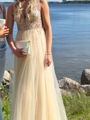 Balklänning - Champagnefärgad  - Elegant långklänning i champagnefärg med glittrig paljettöverdel och v-ringning både fram och baksida. Kjolen är i flera tunna lager. Perfekt för bal eller liknande. 