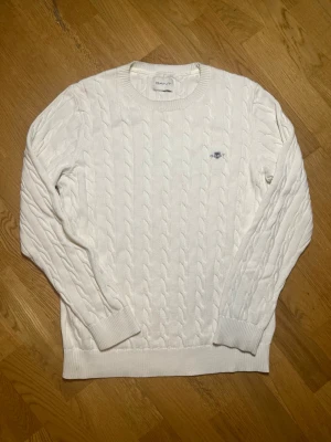 Vit kabelstickad tröja GANT - Säljer denna stilrena kabelstickad vit tröja från GANT med rund halsringning och ribbade muddar. Tröjan har GANT:s klassiska logga broderad på bröstet och är tillverkad i ett mjukt material som passar perfekt till lager på lager. Långärmad och stilren design. Vid frågor, extra bilder eller liknande är det bara att kontakta mig!