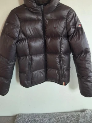 Brun dunjacka från Tommy Hilfiger Denim - Snygg och varm brun dunjacka från Tommy Hilfiger Denim. Jackan har huva, dragkedja framtill, två fickor med dragkedja och logga på ärmen. Passar perfekt till höst och vinter. Innerlapp med detaljerad märkning och storlek medium. Klassisk och tidlös modell.