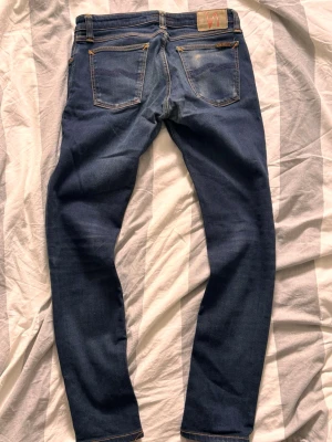  Nudie Jeans slim - Säljer ett par mörkblå jeans från Nudie Jeans med smal passform. Jeansen har kontrastsömmar och är tillverkade i stretchig denim för extra komfort. Perfekta för dig som gillar en clean och stilren look.