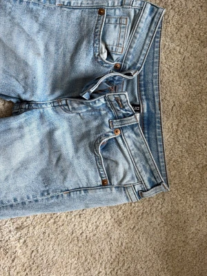 Ljusblå raka jeans med stora bakfickor - Säljer ett par ljusblå jeans med stora bakfickor med knappar. Jeansen har klassisk femficksdesign fram och detaljer i kopparfärg. Materialet är denim i bomull och passformen är avslappnad.