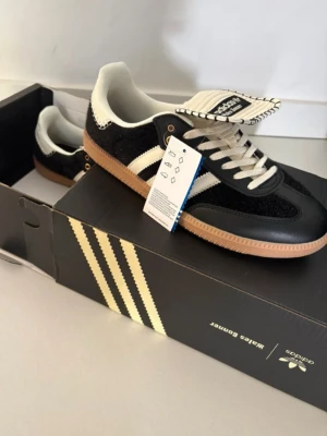 Adidas Wales Bonner svart/vit sneaker - Snygga Adidas Wales Bonner sneakers i svart och vitt med klassiska ränder på sidan. Skorna har en låg silhuett, vita snören och detaljer i både mocka och läder. Yttersulan är i gummi och ger en retrovibe. Perfekta för dig som gillar stilrena och trendiga sneakers.