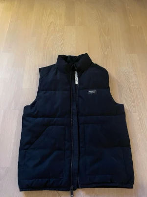Svart dunväst från Abercrombie & Fitch - Stilren svart dunväst från Abercrombie & Fitch med hög krage och dragkedja framtill. Västen har två stora fickor och en diskret logotyp på bröstet. Perfekt för lager-på-lager och ger en clean look till din outfit.