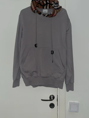 Grå hoodie från Burberry med mönstrad huva - Säljer en grå hoodie från Burberry med klassisk rutmönstrad huva i beige, svart, vitt och rött. Hoodien har dragsko, stor magficka och ribbade muddar. Perfekt för dig som vill ha en stilren men ändå unik look.