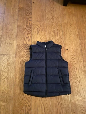 Svart vadderad dunväst - Snygg svart dunväst med quiltad design och dragkedja framtill. Västen har två fickor med dragkedja och hög krage för extra värme. Perfekt att slänga över en hoodie eller tröja för en chill look. Materialet är mjukt och puffigt, vilket ger en modern och sportig vibe.
