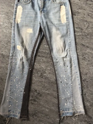 Ljusblå jeans med färgstänk och slitningar - Unika ljusblå jeans med kontrasterande mörka paneler längs benen, slitna detaljer och råa benslut. Jeansen har vita och svarta färgstänk framtill och baktill samt patchade partier. Perfekt för dig som gillar en edgy och kreativ stil.