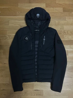 Svart pufferjacka från Sail Racing - Säljer en svart pufferjacka från Sail Racing med huva och dragkedja. Jackan har logga på bröstet och ärmen samt diskreta detaljer. Tillverkad i slitstarkt material som håller värmen under kalla dagar. Perfekt för dig som vill ha en stilren och funktionell vinterjacka.