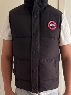 Svart dunväst från Canada Goose - Snygg svart dunväst från Canada Goose med hög krage och tryckknappar framtill. Klassisk patch med logga på bröstet. Västen har två fickor och är perfekt för lager-på-lager under kyliga dagar. Tidlös design som funkar till det mesta.