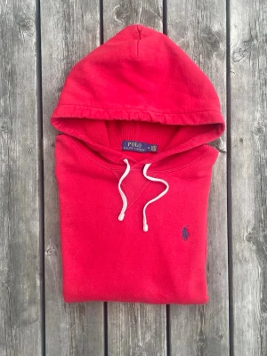 Polo Ralph Lauren Hoodie | Röd - Strl M - Snygg röd hoodie från Polo Ralph Lauren med klassisk blå logga broderad på bröstet. Hoodien har vit dragsko i huvan och är tillverkad i mjukt bomullsmaterial. Perfekt för en avslappnad och stilren look. Fint skick (Obs har en liten imperfektion på ryggen) !