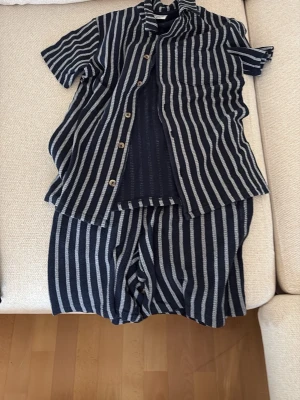 Randigt set med skjorta och shorts - Säljer ett snyggt set med kortärmad skjorta och matchande shorts i marinblått med vita vertikala ränder. Skjortan har knappar framtill och en bröstficka. Perfekt för en avslappnad och trendig look. Materialet känns mjukt och luftigt.