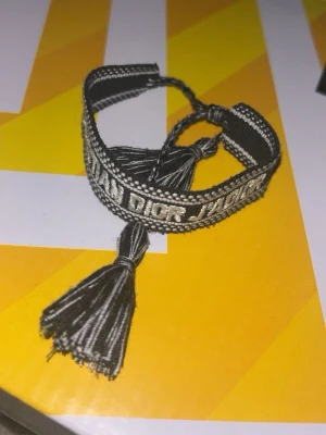 Svart broderat armband med tofsar - Ett snyggt svart armband från Dior med broderad text och dekorativa tofsar i ändarna. Armbandet är justerbart och har en cool, ungdomlig stil med tydliga kontraster mellan svart och vitt. Perfekt för att ge din outfit en exklusiv touch.