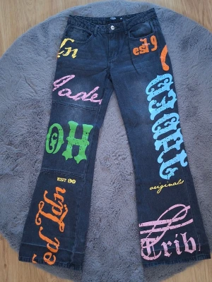 Svarta jeans med färgglada tryck - Unika svarta jeans med utsvängda ben och stora färgglada tryck i orange, rosa, blått och grönt över hela byxorna. Snygga detaljer med olika texter och grafiska element både fram och bak. Perfekta för dig som vill sticka ut med din stil. Har lite märken längst ner från att ha varit upplagda, men syns inte när de sitter på.