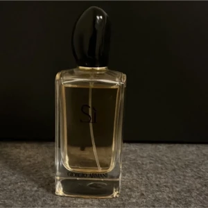 Giorgio Armani Sì parfym 50ml - Elegant parfymflaska från Giorgio Armani Sì med genomskinligt glas och svart lock. Flaskan har en stilren, fyrkantig form och innehåller en ljusgul vätska. Perfekt för dig som gillar exklusiva dofter och snygg design på parfymhyllan.