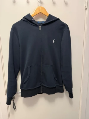 Mörkblå hoodie från Polo Ralph Lauren - Snygg mörkblå hoodie från Polo Ralph Lauren med dragkedja framtill och klassisk vit logga på bröstet. Hoodien har huva med dragsko och två fickor framtill. Tillverkad i mjukt bomullsmaterial som känns skönt mot huden.