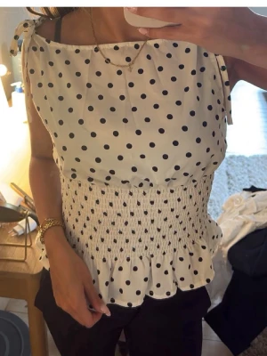 Prickig vit blus med knytband - Söt vit blus med svarta prickar och knytband på axlarna. Toppen har smockad midja och volangkant nedtill för en feminin look. Perfekt till sommaren och lätt att matcha med både jeans och kjol.