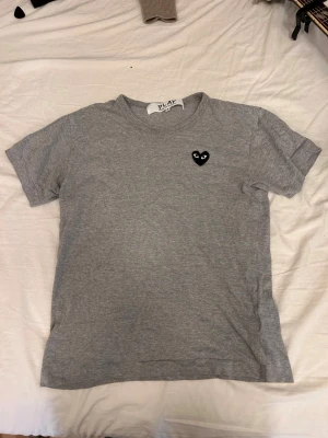 Grå t-shirt Comme des Garçons Play - Snygg grå t-shirt från Comme des Garçons Play med det ikoniska svarta hjärta-logotypen på bröstet. Klassisk rund hals och korta ärmar. Tillverkad i mjuk bomull för en bekväm känsla. Perfekt till jeans eller shorts.
