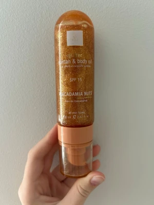 Glittrig sol- och kroppsolja SPF 15 - Glittrig sol- och kroppsolja med SPF 15 och doft/innehåller macadamianötter. Kommer i en genomskinlig pumpflaska med guldigt skimmer som ger huden extra glow. Passar alla hudtyper och är enkel att applicera för en snygg finish.