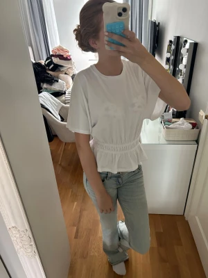 Vit topp med volang och resårmidja - Säljer en söt vit topp med rund hals, korta ärmar och resår i midjan som ger en snygg volangkant. Perfekt till jeans eller kjol för en fräsch look. Materialet känns mjukt och luftigt, troligen bomull eller polyester.
