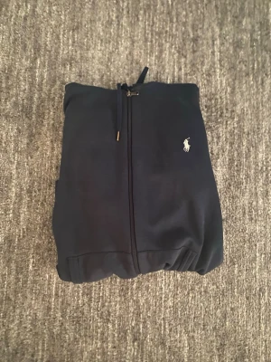 Ralph Lauren hoodie - Mörkblå hoodie med dragkedja från Ralph Lauren. Klassisk design med huva, fickor framtill och den ikoniska vita loggan broderad på bröstet. Perfekt för en chill och stilren look. Mjuk bomullskänsla och ribbade muddar.