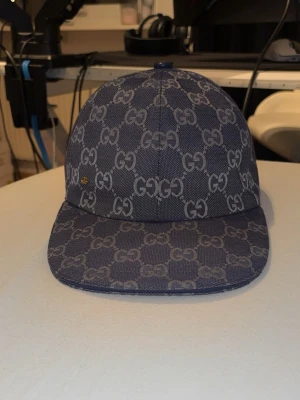 Blå keps med GG mönster  - Snygg blå keps med det klassiska GG-monogrammet i ett rutigt mönster över hela kepsen. Kepsen har böjd skärm och justerbar rem bak. Tillverkad i ett slitstarkt tyg som ger en exklusiv känsla.