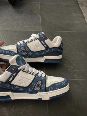 Louis Vuitton blåvita sneakers - Säljer ett par riktigt snygga Louis Vuitton sneakers i blått och vitt. Skorna har klassiskt monogrammönster i denimtyg, vita läderdetaljer och robust sula. Perfekta för dig som vill sticka ut med exklusiv streetwear och unik design.
