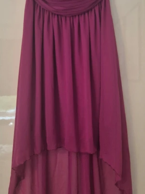 Långkjol i mörkrosa med slits - Säljer en snygg mörkrosa långkjol med asymmetrisk fåll och slits. Kjolen har ett tunt, flytande material och dragkedja bak. Perfekt för dig som vill ha en statement piece i garderoben.