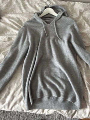 Grå hoodie i mjukt material - En stilren grå hoodie med huva och dragsnören. Tillverkad i ett mjukt, stickat material som känns skönt mot huden. Hoodien har ribbade muddar vid ärmslut och nederkant samt en avslappnad passform. Perfekt för en chill och avslappnad look.