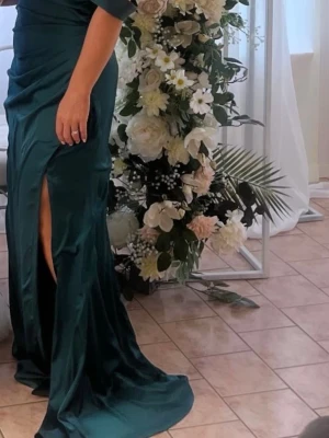 Mörkgrön lång satinklänning - Snygg mörkgrön långklänning i glansigt satintyg med breda axelband och draperad överdel. Klänningen har en hög slits framtill och figurnära passform som ger en elegant siluett. Perfekt för bal eller festliga tillfällen.