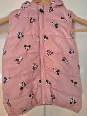 Rosa dunväst med Disney-mönster - Söt rosa dunväst med huva och dragkedja framtill. Västen är täckt av små tryck med Musse och Mimmi Pigg i svart och vitt. Perfekt för dig som gillar Disney och vill ha en lekfull stil. Materialet är quiltat och ger en puffig känsla.i storlek 86
