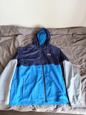 Vindjacka Under Armour - Snygg vindjacka från Under Armour i blått och marinblått med ljusgrå ärmar. Jackan har huva, dragkedja framtill och två sidofickor. Perfekt för blåsiga dagar och sportiga outfits.
