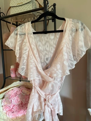 Ljusrosa omlottopp med volang - Supersöt ljusrosa omlottopp med volangdetaljer och knyt i midjan. Tunn, luftig och med ett diskret leopardmönster. Perfekt för dig som vill ha en feminin och trendig look. Djup v-ringning och korta, fladdriga ärmar.