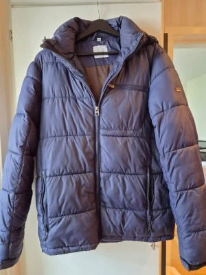 Blå pufferjacka från Erla of Sweden - Snygg blå pufferjacka från Erla of Sweden med dragkedja framtill, två sidofickor med dragkedja och en bröstficka. Jackan har hög krage och quiltad design som håller värmen. Perfekt för kalla vinterdagar.