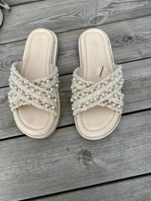 Beige sandaler med pärldetaljer - Säljer ett par snygga beige sandaler med korsade band dekorerade med pärlor och fransiga kanter. Sandalerna har en mjuk sula och öppen tå, perfekta för sommarens alla tillfällen. Materialet är textil och syntet.